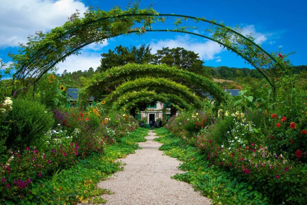 Stone pathway with wildflowers and plant arches leading to a house with an open front door.